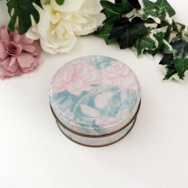 Boîte ronde en métal au décor pastel de pivoines roses et feuillages bleus, posée sur un fond clair avec des fleurs en arrière-plan.
