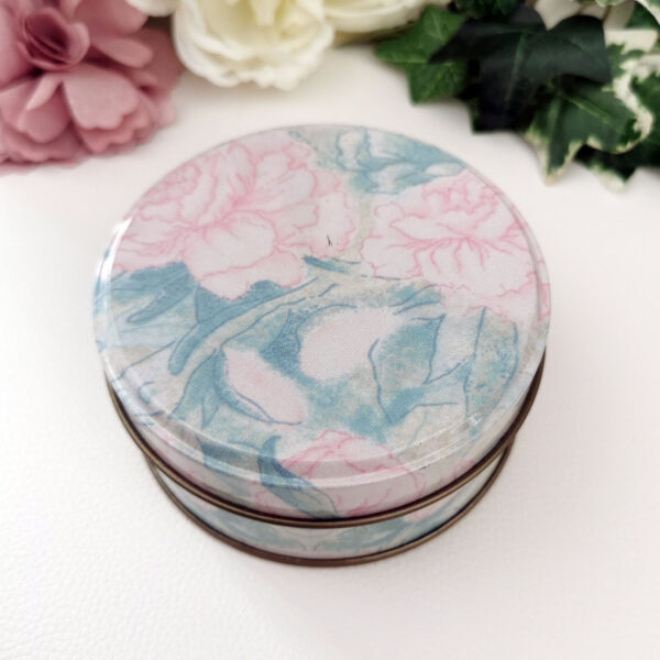 Boîte ronde en métal au décor pastel de pivoines roses et feuillages bleus, posée sur un fond clair avec des fleurs en arrière-plan.
