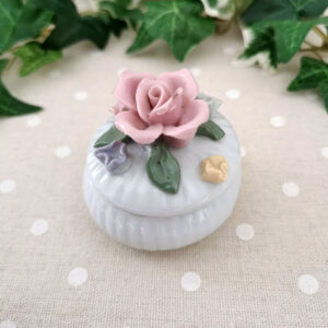 Mini-boîte ronde en porcelaine blanche décorée de fleurs pastel en relief, posée sur un tissu à pois.