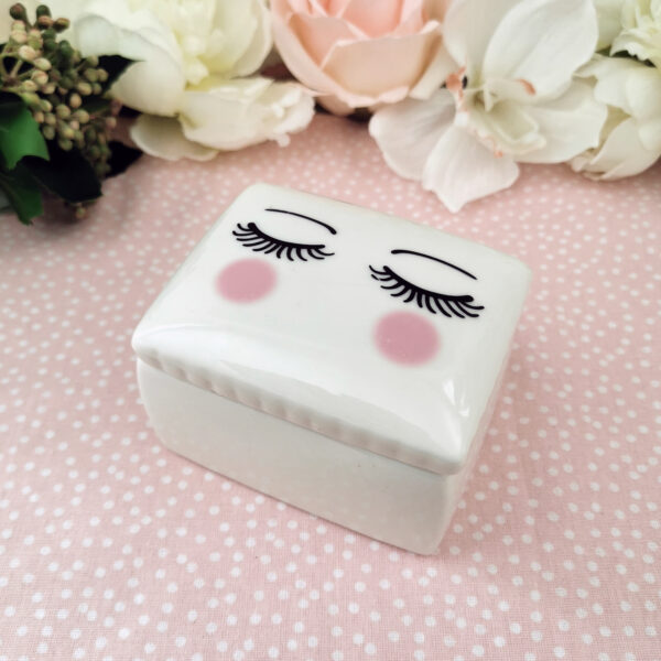 Petite boîte en céramique blanche décorée d’un visage stylisé aux cils fermés et joues roses